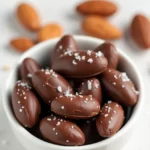 Homemade chocolate covered almonds with glossy dark chocolate coating and sea salt flakes arranged on a white ceramic plate, showcasing both whole pieces and cross-sections revealing the roasted almond inside.